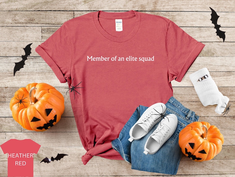 May include: A red heather t-shirt with the text "Member of an elite squad" printed in white on the front. The shirt is laid out on a wooden surface with a pair of white sneakers, blue jeans, and a white sock with a black spider on it.
