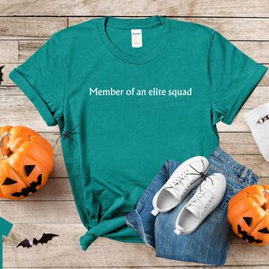 May include: Teal t-shirt with the text "Member of an elite squad" printed in white. The shirt is laying on a wooden surface with a pair of white sneakers and a pair of jeans.