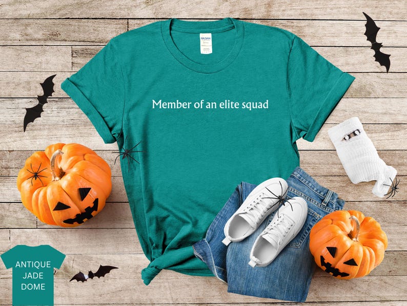 May include: Teal t-shirt with the text "Member of an elite squad" printed in white. The shirt is laying on a wooden surface with a pair of white sneakers and a pair of jeans.