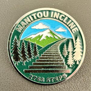 May include: A round metal pin featuring a green and blue enamel design of the Manitou Incline. The image depicts a mountain with white snow, pine trees, and a long staircase leading upward. The text reads MANITOU INCLINE at the top and 2768 STEPS at the bottom.