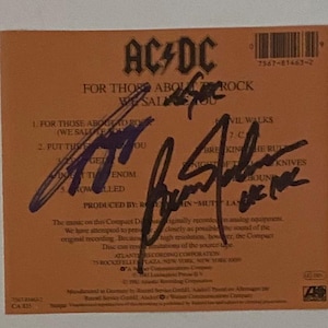 May include: A close-up of the back of a CD case for the album "For Those About to Rock (We Salute You)" by AC/DC. The case is orange and features the band's logo and a list of songs. The case is signed by the band members.