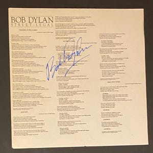Pode incluir: Uma folha de papel impressa em preto e branco com as letras de duas músicas de Bob Dylan: "Changing of the Guards" e "Baby Stop Crying". As letras são escritas à mão em tinta azul. A folha é assinada por Bob Dylan em tinta azul.