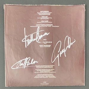 May include: A vintage vinyl record album cover with signatures. The cover is a muted brown color with white text and signatures. The text includes the names of the band members, song titles, and production credits. The signatures are large and prominent.