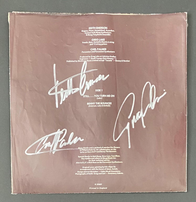 May include: A vintage vinyl record album cover with signatures. The cover is a muted brown color with white text and signatures. The text includes the names of the band members, song titles, and production credits. The signatures are large and prominent.