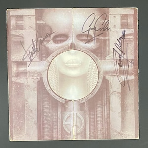 May include: Album cover with a surreal, sepia-toned image featuring a human face within a circular frame, surrounded by mechanical structures. The cover is signed by multiple people. The overall theme is science fiction.