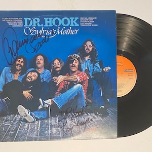 Puede incluir: Un disco de vinilo negro con una portada de álbum azul y blanca con una banda llamada Dr. Hook. La portada del álbum tiene el título "Subria's Mother" y presenta una foto de la banda con los miembros de la banda sentados en un suelo de madera. La portada del álbum está firmada por los miembros de la banda.