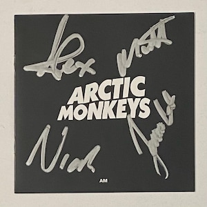 May include: A black square with the band name "Arctic Monkeys" in white, and four silver signatures. The initials "AM" are in the bottom right corner. The signatures are likely from the band members.