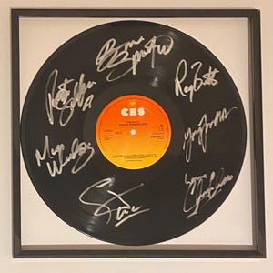 以下が含まれることがあります： 署名入りの黒いビニールレコードが額装されています。レコードの中央にはオレンジと黄色のCBSレーベルがあります。署名は銀色のインクです。レコードは黒いフレームに飾られています。