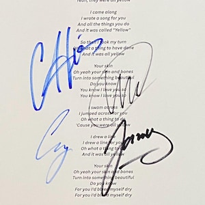 Puede incluir: Una hoja de papel blanco con la letra de la canción Yellow impresa en negro, incluyendo líneas como I came along, I wrote a song for you y And it was all yellow. El documento cuenta con cuatro firmas manuscritas distintas en tinta azul y negra.