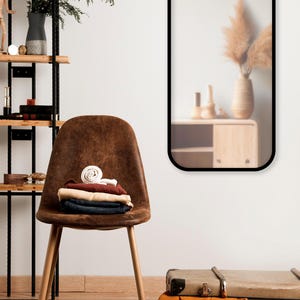 Peut inclure: Une chaise marron avec des vêtements pliés, des chaussures noires et une valise. Un miroir encadré de noir reflète un vase avec des plantes séchées. Une étagère avec des objets décoratifs est visible.