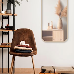 Op de afbeelding: Een bruine stoel met houten poten houdt een stapel opgevouwen kleding vast. Een rechthoekige spiegel met afgeronde hoeken hangt aan de muur. Een paar zwarte schoenen staat op de vloer, in de buurt van een koffer en een oranje deken.