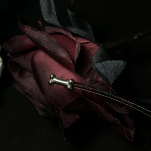 May include: A deep burgundy fabric rose with black fabric leaves. A silver-coloured bone-shaped charm is attached to the rose. The background is black, creating a dramatic contrast.