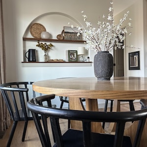 K&ouml;nnte beinhalten: Ein Esszimmer mit einem runden Holztisch und schwarzen St&uuml;hlen. Eine dunkelgraue Vase enth&auml;lt wei&szlig;e Bl&uuml;tenzweige. Ein eingebautes Regal zeigt dekorative Gegenst&auml;nde, darunter einen geflochtenen Korb und gerahmte Kunstwerke. &Uuml;ber dem Tisch h&auml;ngt ein moderner Kronleuchter.