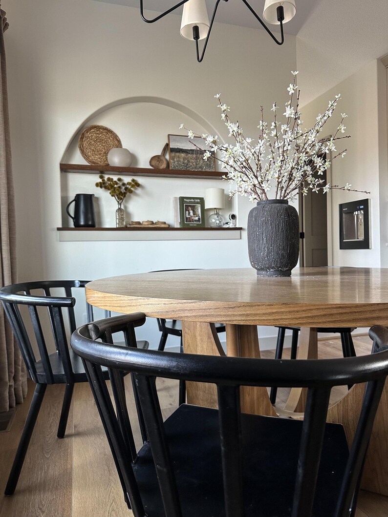 K&ouml;nnte beinhalten: Ein Esszimmer mit einem runden Holztisch und schwarzen St&uuml;hlen. Eine dunkelgraue Vase enth&auml;lt wei&szlig;e Bl&uuml;tenzweige. Ein eingebautes Regal zeigt dekorative Gegenst&auml;nde, darunter einen geflochtenen Korb und gerahmte Kunstwerke. &Uuml;ber dem Tisch h&auml;ngt ein moderner Kronleuchter.
