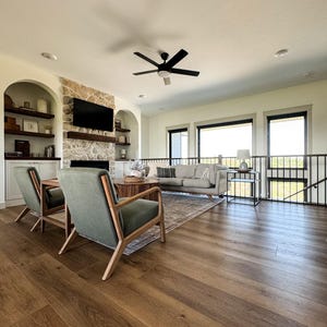 K&ouml;nnte beinhalten: Ein modernes Wohnzimmer mit Holzb&ouml;den, zwei gr&uuml;nen Sesseln und einem grauen Sofa. Ein schwarzer Deckenventilator h&auml;ngt von der Decke. Ein Steinkamin mit Fernseher und Einbauregalen ist im Hintergrund zu sehen. Gro&szlig;e Fenster bieten einen Blick nach drau&szlig;en.