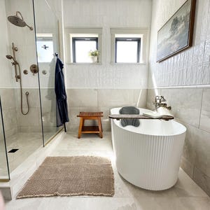 K&ouml;nnte beinhalten: Ein modernes Badezimmer mit einer wei&szlig;en, geriffelten Badewanne, einem Holzschemel und einem gewebten beigefarbenen Teppich. Die Dusche hat eine Glast&uuml;r und bronzefarbene Armaturen. Ein gerahmtes Landschaftsbild h&auml;ngt an der Wand, und zwei Fenster spenden nat&uuml;rliches Licht.