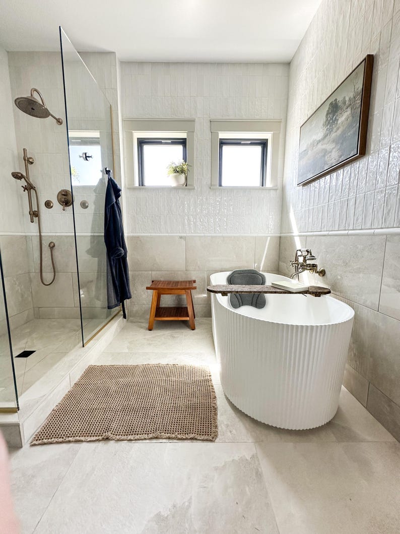 K&ouml;nnte beinhalten: Ein modernes Badezimmer mit einer wei&szlig;en, geriffelten Badewanne, einem Holzschemel und einem gewebten beigefarbenen Teppich. Die Dusche hat eine Glast&uuml;r und bronzefarbene Armaturen. Ein gerahmtes Landschaftsbild h&auml;ngt an der Wand, und zwei Fenster spenden nat&uuml;rliches Licht.