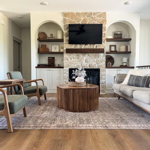 K&ouml;nnte beinhalten: Ein Wohnzimmer mit Steinkamin, schwarzem Fernseher und Einbauregalen. Ein runder Couchtisch aus Holz steht auf einem gemusterten Teppich. Zwei gr&uuml;ne Sessel und ein graues Sofa vervollst&auml;ndigen den Raum.