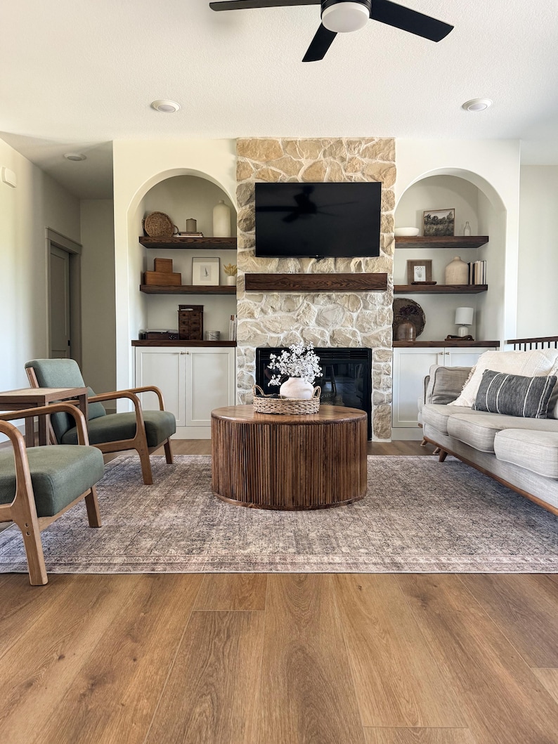 K&ouml;nnte beinhalten: Ein Wohnzimmer mit Steinkamin, schwarzem Fernseher und Einbauregalen. Ein runder Couchtisch aus Holz steht auf einem gemusterten Teppich. Zwei gr&uuml;ne Sessel und ein graues Sofa vervollst&auml;ndigen den Raum.