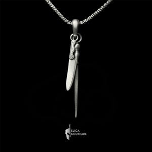 May include: A silver-toned pendant necklace featuring a stylized knife and a bundle of thin, parallel lines, possibly representing a sword or a similar weapon. The pendant hangs from a delicate chain against a black background. The brand name "ELICA BOUTIQUE" is at the bottom.