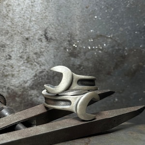 Könnte beinhalten: Zwei silberne Ringe in Form eines Schraubenschlüssels, die übereinander gestapelt sind. Die Ringe liegen auf einer Metalloberfläche.