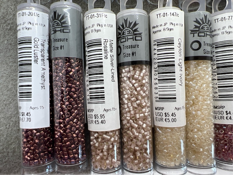 May include: Six tubes of TOHO Treasure seed beads in various colours, including gold luster, transparent amethyst, matte silver-lined Rosaline, and Ceylon light ivory. Each tube is labelled with size #1 and contains approximately 8.9 grams of beads.