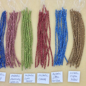 May include: Assortment of metallic beads in various colors, including navy, pink, green, red, blue, and gold. The beads are arranged in rows on a light brown surface with labels identifying each color.