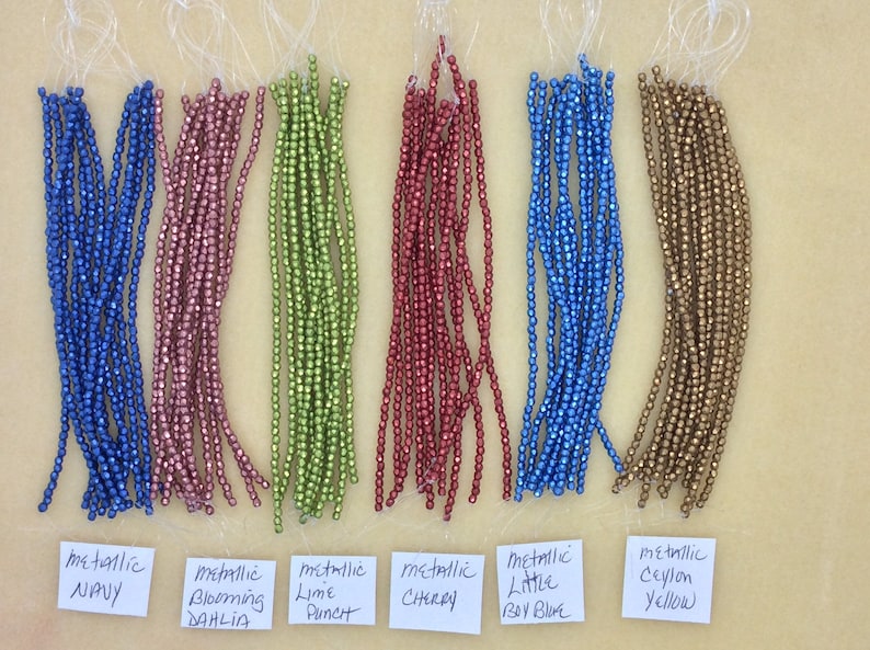 May include: Assortment of metallic beads in various colors, including navy, pink, green, red, blue, and gold. The beads are arranged in rows on a light brown surface with labels identifying each color.