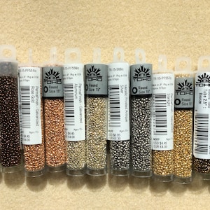 May include: Ten small tubes of TOHO seed beads in various metallic finishes, including rose gold, silver, and bronze. Each tube is labeled with the bead size, color, and material.