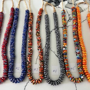 Puede incluir: Colección de seis collares de cuentas de varios colores. Los collares están ensartados en un cordón natural y presentan una variedad de patrones de cuentas, incluyendo rojo, azul, naranja y negro. Cada collar está etiquetado con un número.