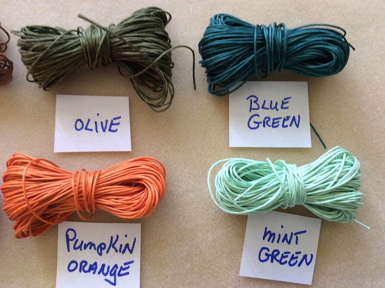 May include: Five spools of waxed cotton cord in different colors: olive green, blue green, pumpkin orange, mint green, and brown. The spools are labeled with their colors.