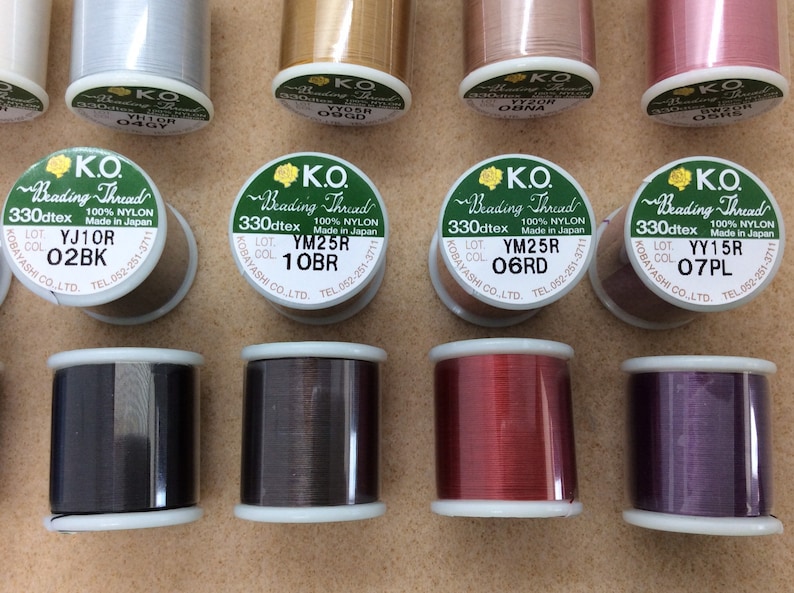 May include: Close-up of ten spools of thread for beading. The spools are labelled with the brand "K.O. Beading Thread" and the colour and lot numbers. The colours of the thread are silver, gold, pink, red, black, brown, red, and purple.