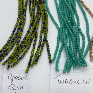 May include: Two strands of beads, one in opaque olive green with iridescent accents and the other in turquoise. The beads are labeled "Opaque Olive Vitral" and "Turquoise".