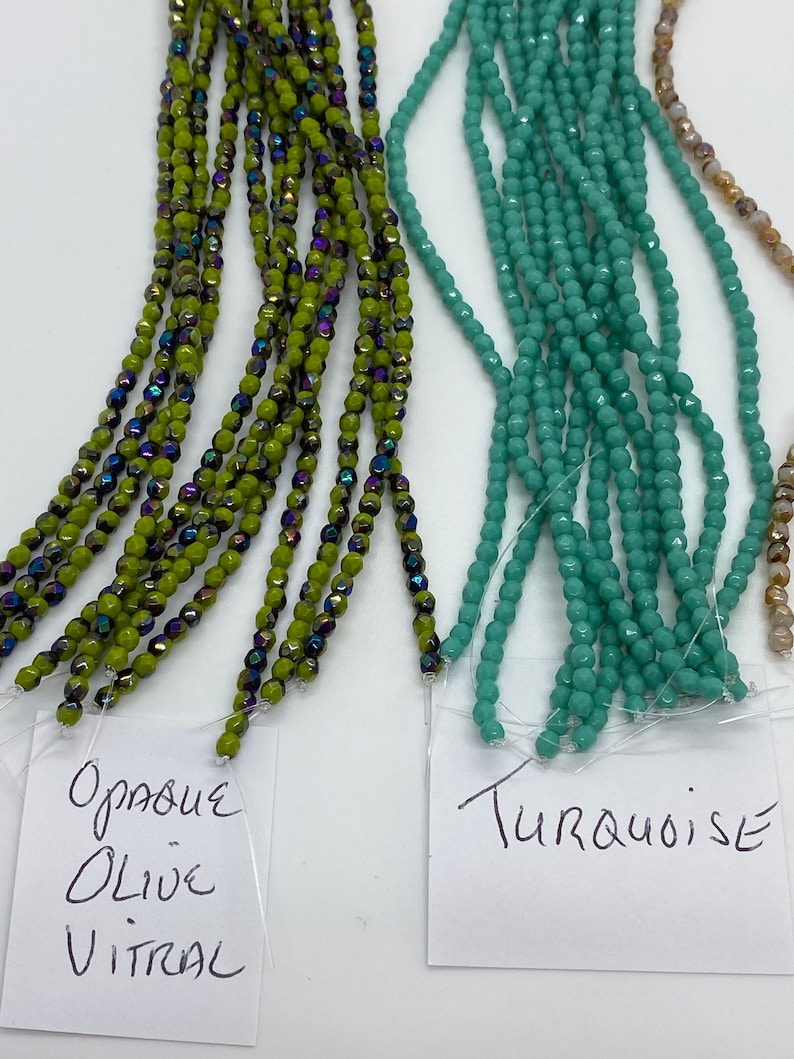 May include: Two strands of beads, one in opaque olive green with iridescent accents and the other in turquoise. The beads are labeled "Opaque Olive Vitral" and "Turquoise".