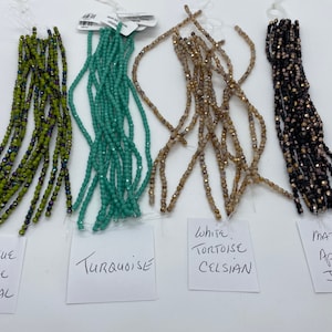 May include: Five strands of glass beads in different colors and finishes. From left to right: Opaque Olive Vitrail, Turquoise, White Tortoise Celsian, Matte Apollo Jet.