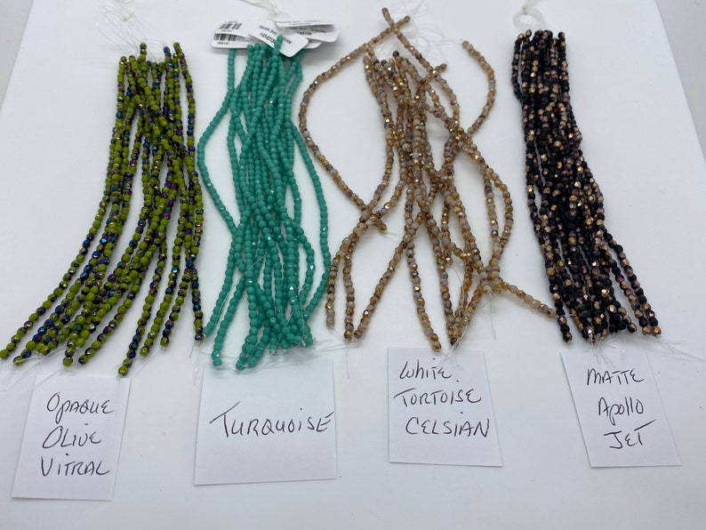 May include: Five strands of glass beads in different colors and finishes. From left to right: Opaque Olive Vitrail, Turquoise, White Tortoise Celsian, Matte Apollo Jet.