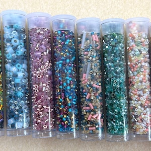May include: Eight clear plastic tubes filled with colorful beads. The beads are in various colors, including green, blue, purple, and gold. The tubes are arranged side by side, each with a clear plastic lid. The beads appear to be for jewelry making or other crafts.