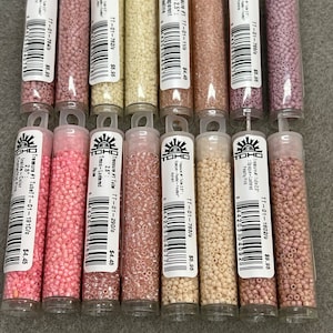 May include: A selection of Toho Treasure seed beads in shades of pink, peach, and cream, presented in clear plastic tubes. Each tube is labelled with product details, including colour and size, and the price.