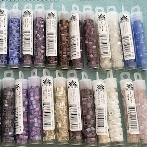 May include: A collection of 20 small tubes of TOHO seed beads in various colors, including purple, white, silver, and blue. Each tube is labeled with the bead size, color, and price. The tubes are arranged in two rows of ten.