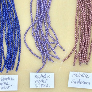 May include: Three strands of metallic beads in different colors: super violet, ballet slipper, and butternum. The beads are small and have a faceted shape.