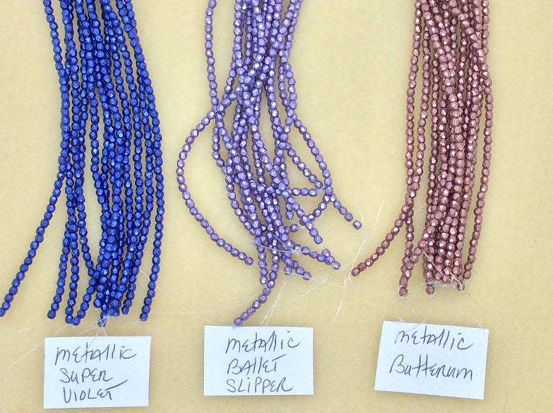 May include: Three strands of metallic beads in different colors: super violet, ballet slipper, and butternum. The beads are small and have a faceted shape.