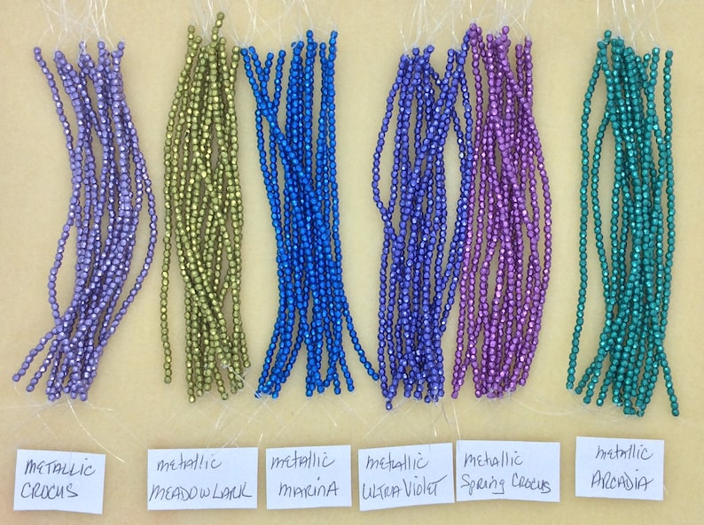 May include: Six strands of metallic beads in various colors, including purple, green, blue, and teal. The beads are labeled with their color names: Metallic Crocus, Meadow Lark, Marina, Ultra Violet, Spring Crocus, and Arcadia.