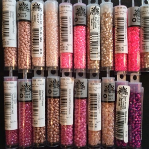 May include: A collection of 14 small tubes of TOHO brand round seed beads in various colors, including pink, beige, and purple. Each tube has a label with the bead size, color, and product code.