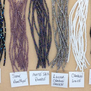 May include: Six strands of glass beads in various colors and finishes, including jet AB, stone amethyst, matte iris purple, luster opaque green, opaque white, and jet black. The beads are arranged in a row on a brown surface with labels underneath each strand.