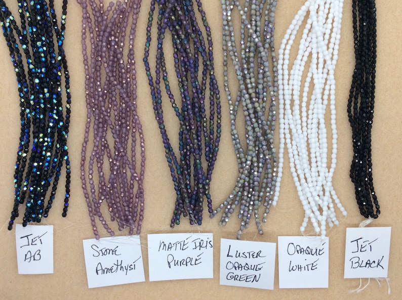 May include: Six strands of glass beads in various colors and finishes, including jet AB, stone amethyst, matte iris purple, luster opaque green, opaque white, and jet black. The beads are arranged in a row on a brown surface with labels underneath each strand.