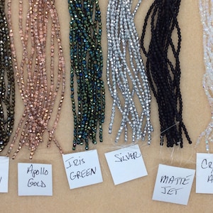 May include: Six strands of faceted glass beads in various colors, including brown, gold, green, silver, black, and clear. The beads are labeled with their names: Iris Brown, Apollo Gold, Iris Green, Silver, Matte Jet, and Crystal AB.