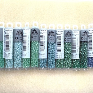 May include: A collection of small, colorful glass beads in clear tubes. The beads are in shades of green, blue, and silver. Each tube has a label with the brand name "TOHO" and product information. The beads are size 8.