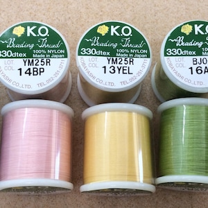 May include: Four spools of nylon beading thread in different colours: pink, yellow, green, and blue. Each spool has a label with the text "K.O. Beading Thread 330dtex 100% Nylon Made in Japan" and a lot number and colour code.