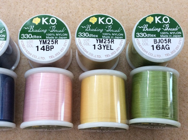 May include: Four spools of nylon beading thread in different colours: pink, yellow, green, and blue. Each spool has a label with the text "K.O. Beading Thread 330dtex 100% Nylon Made in Japan" and a lot number and colour code.