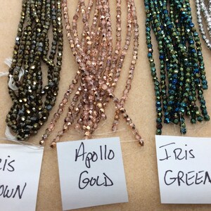 May include: Four strands of glass beads in different colors. The beads are iridescent and have a faceted finish. The strands are labeled "Iris Brown", "Apollo Gold", "Iris Green", and "Silver".